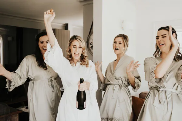Bride celebrating with bridesmaids while opening a bottle of champagne.